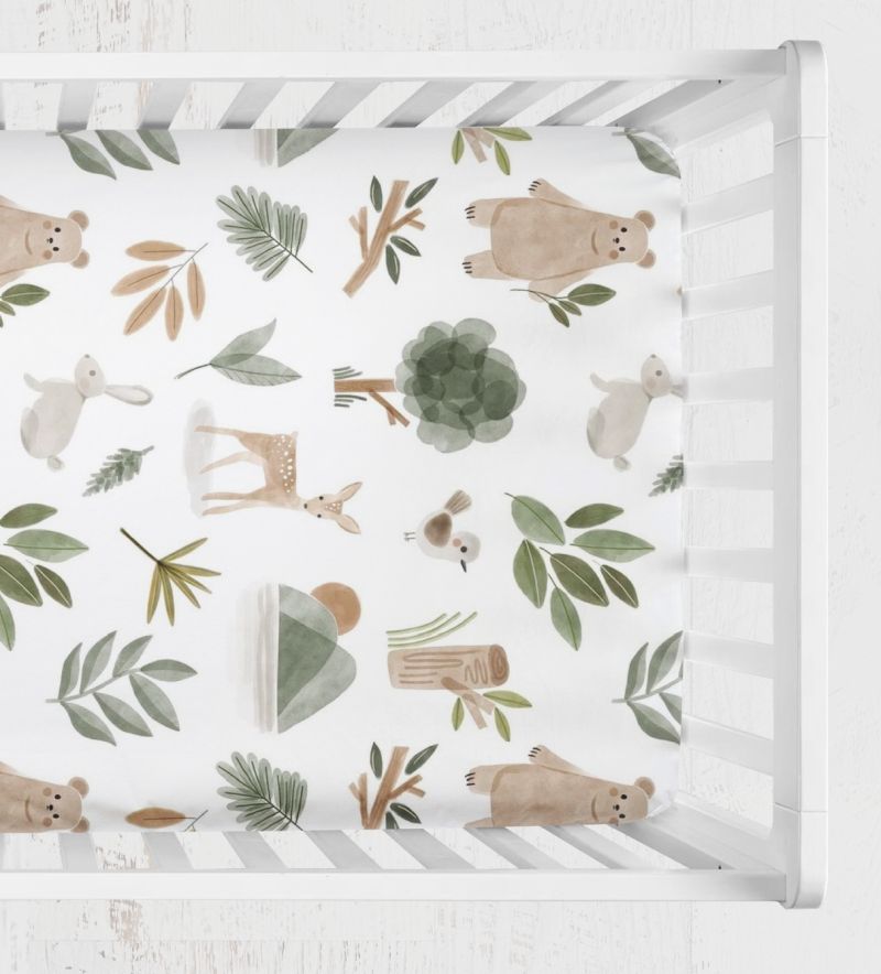 A top-down view of a crib sheet with a woodland pattern of bears, rabbits, deer, leaves, and trees in muted green, brown, and beige tones. woodland animals crib sheet - The Creative Raccoon
