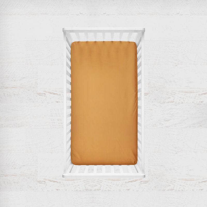 (Yarrow) Mustard yellow fitted sheet on a white crib with a light gray background. The Creative Raccoon